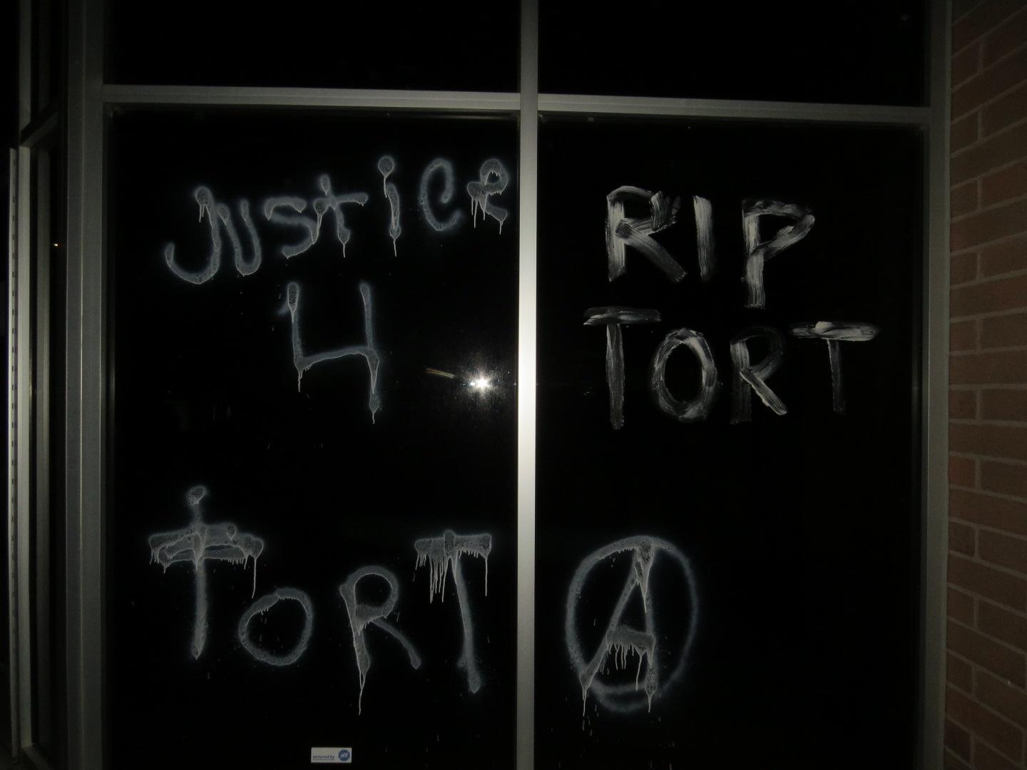 Office building windows, vandalized with acid-etching paint, read "Justice 4 Tort", "RIP TORT" and have a circle-a