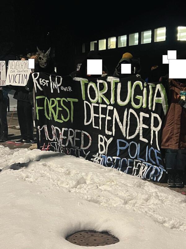 Masked demonstrators on a snowy sidewalk hold a hand-painted banner reading "Rest In Power Tortugita, Forest Defender, Murdered By Police, #stopcopcity #justicefortort"
