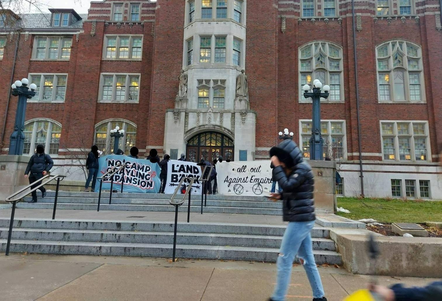 Rally-goers hold banners reading "No Camp Grayling Expansion", "Land Back", and "All Out Against Empire" in front of the Michigan Union building