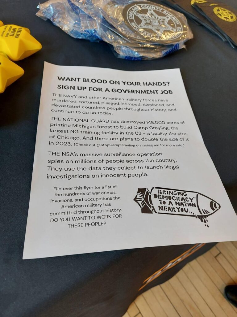 A flyer distributed by protestors lies on the recruiting table for the Oakland County Sheriff. The flyer reads: "WANT BLOOD ON YOUR HANDS? SIGN UP FOR A GOVERNMENT JOB The Navy and other American military forces have murdered, tortured, pillaged, bombed, displaced, and devastated countless people throughout history, and continue to do so today. The National Guard has destroyed 148,000 acres of pristine Michigan forest to build Camp Grayling, the largest NG training facility in the US - a facility the size of Chicago. And there are plans to double the size of it in 2023. (Check out @StopCampGrayling on Instagram for more details) The NSA's massive surveillance operation spies on millions of people across the country. They use the data they collect to launch illegal investigations on innocent people. Flip over this flyer for a list of the hundreds of war crimes, invasions, and occupations the American military has committed throughout history. DO YOU WANT TO WORK FOR THESE PEOPLE?"