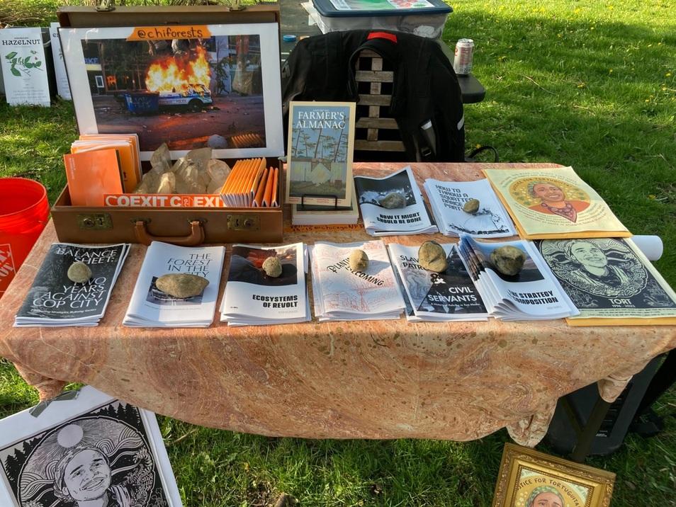 A table with a tablecloth in a park has a number of zines and other free literature on it, as well as a framed photo of a burning cop car, and posters memorializing Tortuguita.