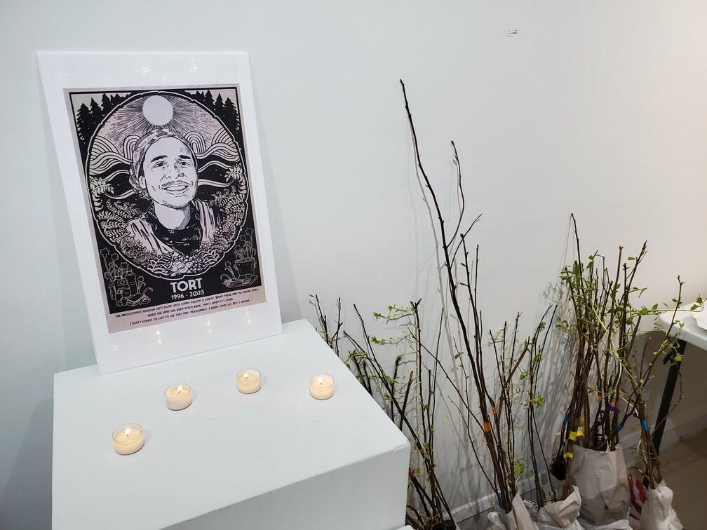 A framed poster memorializing Tortuguita, a forest defender murdered by police while they were protecting the Weelaunee forest in Atlanta, sits on an end table with some small candles. Beside the table are some very young trees in plastic bags with color-coded ribbons, ready to be given away.
