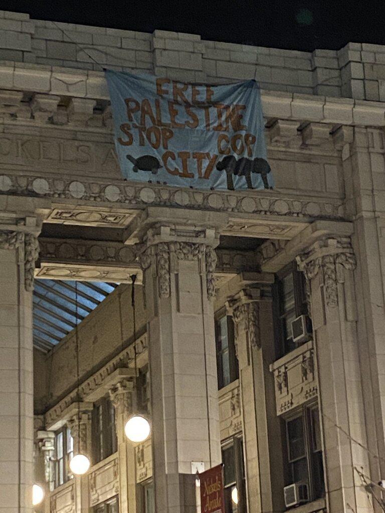 A banner hanging from a roof in downtown Ann Arbor which reads "Free Palestine, Stop Cop City"