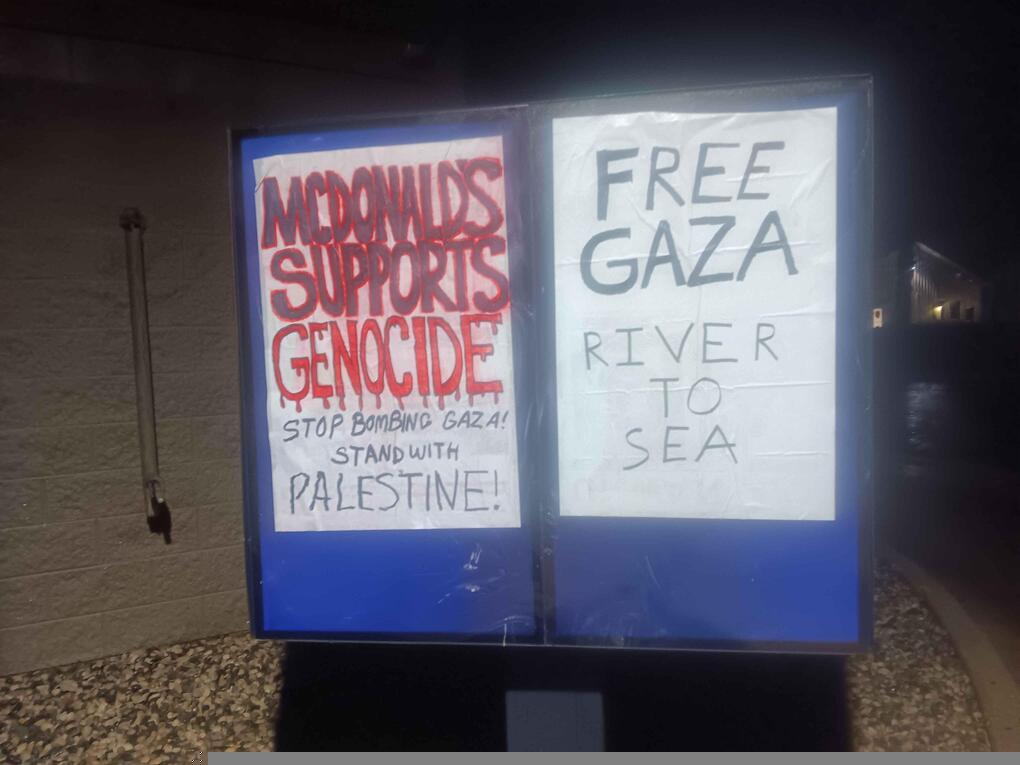 A McDonald's drive-through menu is covered with wheatpasted posters reading "McDonald's Supports Genocide", "Free Gaza", "River to Sea", "Stop Bombing Gaza", and "Stand with Palestine!"