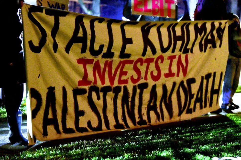 Protestors hold a banner reading "Stacie Kuhlman Invests in Palestinian Death"