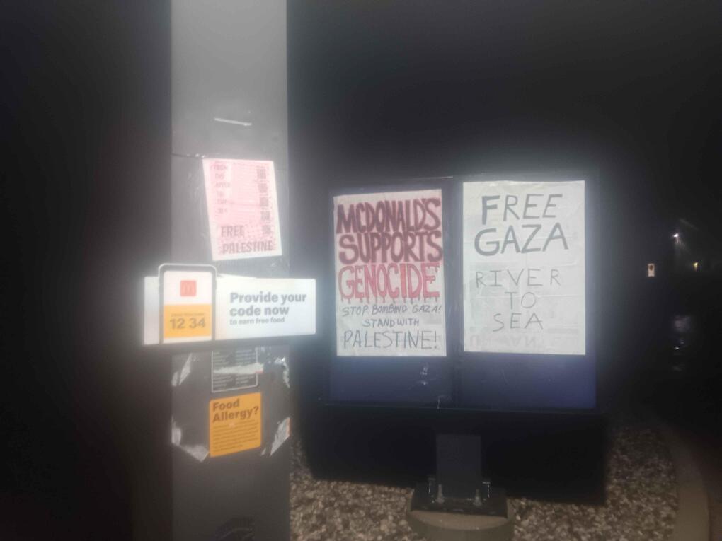 A McDonald's drive-through speaker is covered with a poster reading "Free Palestine". In the background is a drive-through menu covered with posters reading "McDonald's Supports Genocide" and "Free Gaza", with other slogans.