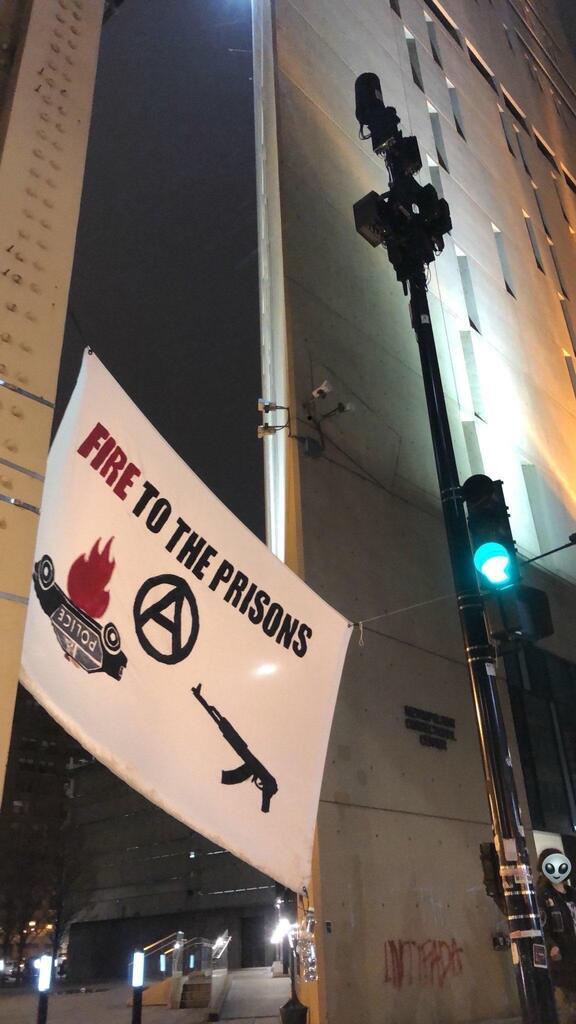 A banner reading "fire to the prisons" with images of a burning cop car, ak-47, and circle-a is hung from a steel girder and a traffic light near a building.