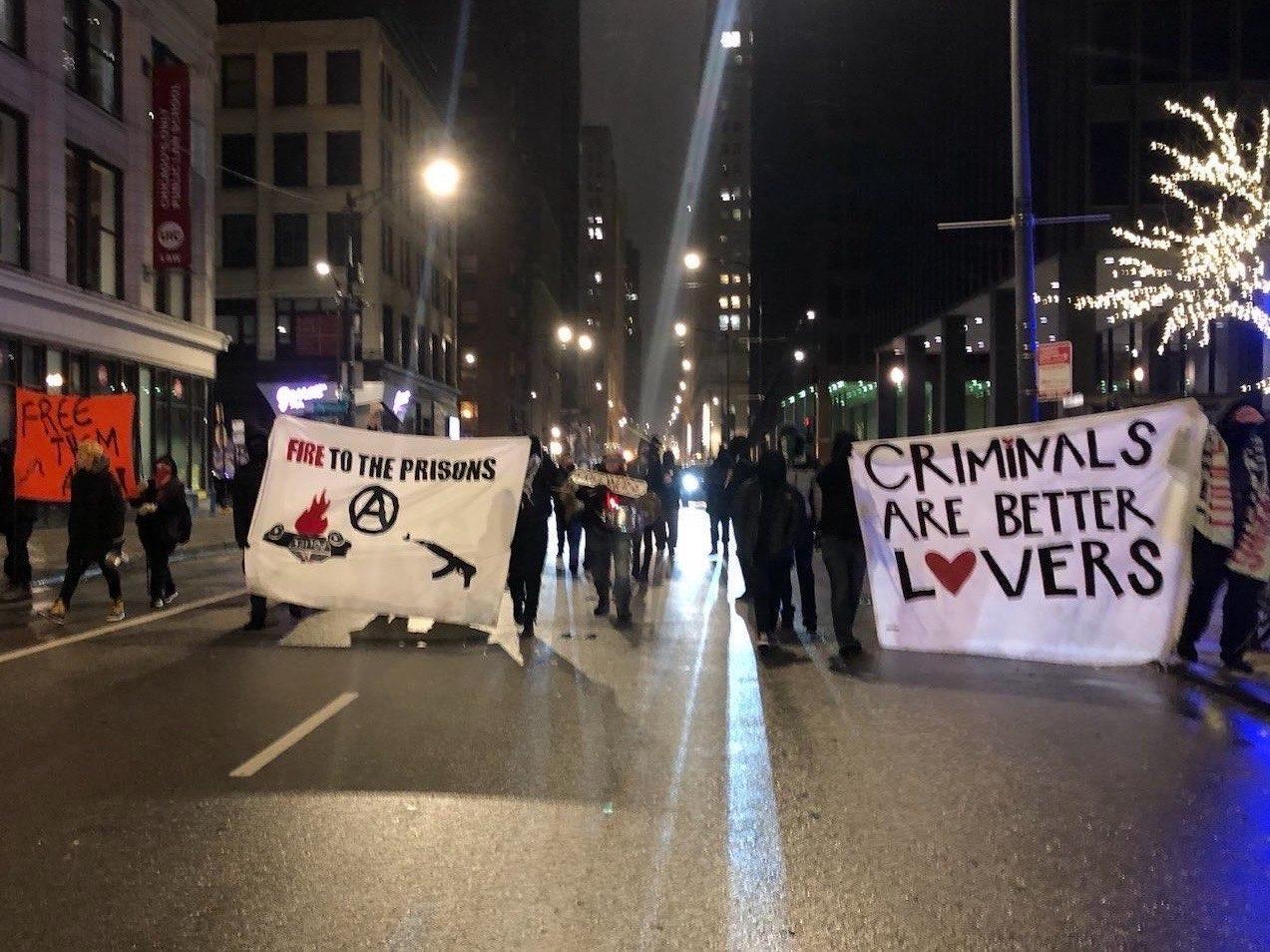 A crowd marches in the street holding banners reading "criminals are better lovers" and "fire to the prisons" with images of a burning cop car, ak-47, and circle-a