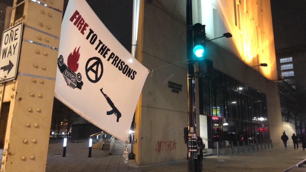 A banner reading "fire to the prisons" with images of a burning cop car, ak-47, and circle-a is hung from a steel girder and a traffic light near a building.