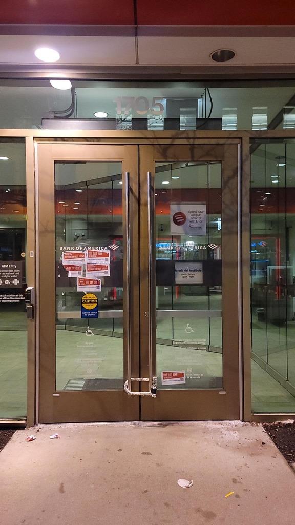 The front doors of a Bank of America branch are locked together with a bicycle u-lock, with some flyers wheat-pasted to the front doors.