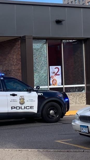 A bank facade has a broken window. A police cruiser is parked in front of it.