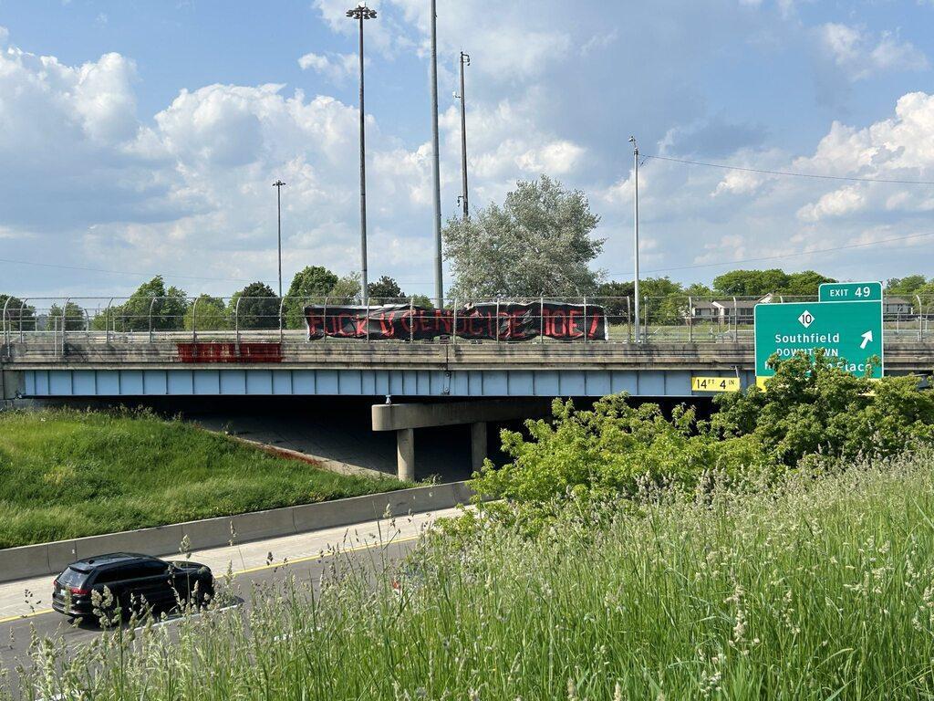 A large black banner on an interstate overpass reads "FUCK U GENOCIDE JOE!" in red text. The interstate below the overpass is visible along with a passing car, and a sign for Exit 49 for M-10 towards Southfield and Downtown