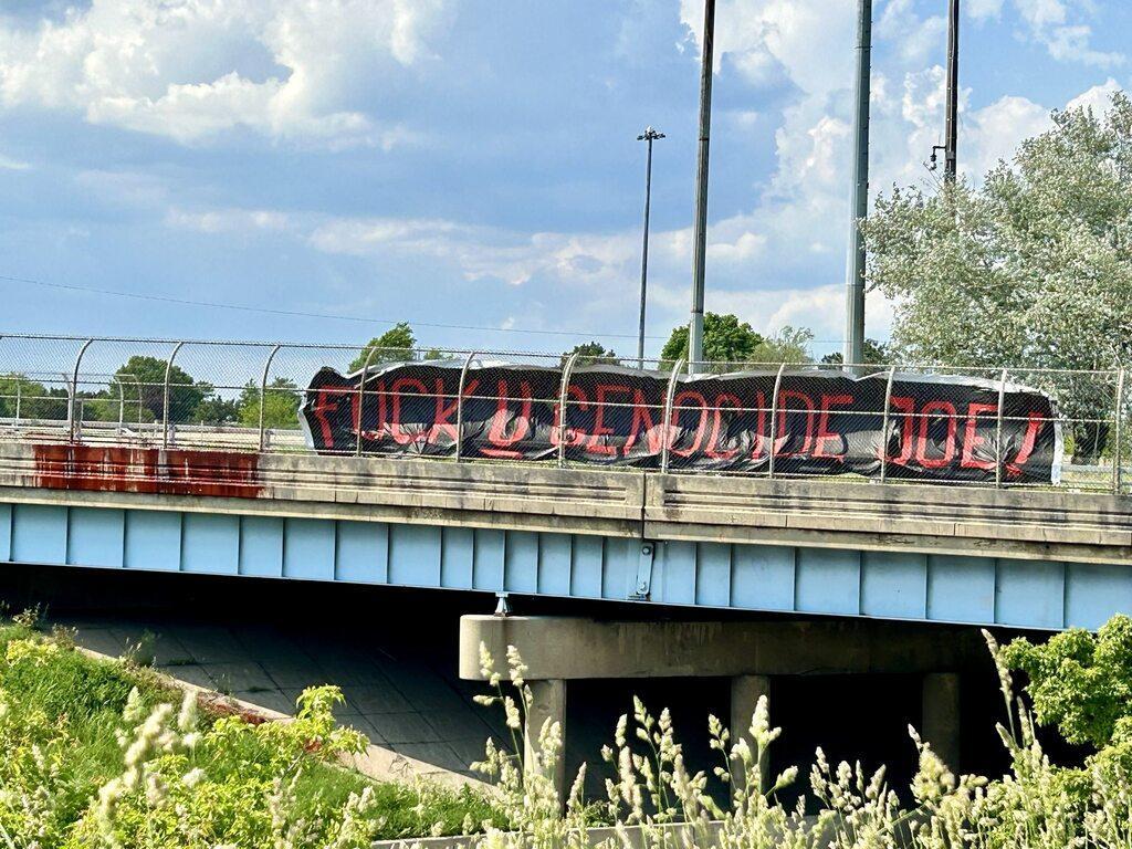 A large black banner on an interstate overpass reads "FUCK U GENOCIDE JOE!" in red text