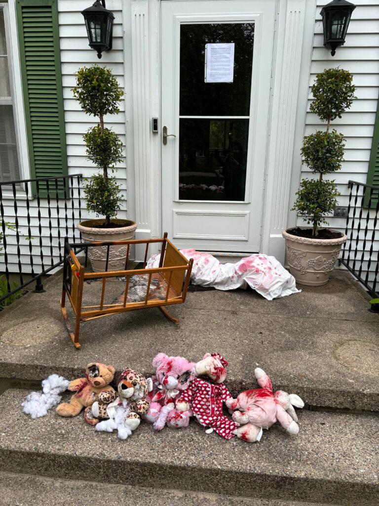 The front porch at a University of Michigan Regent's house has a printed message taped to the front door and props for the home demo. A broken crib, stuffed animals with fake blood, and a fake body in a burial cloth represent Palestinians killed by the Israeli military.