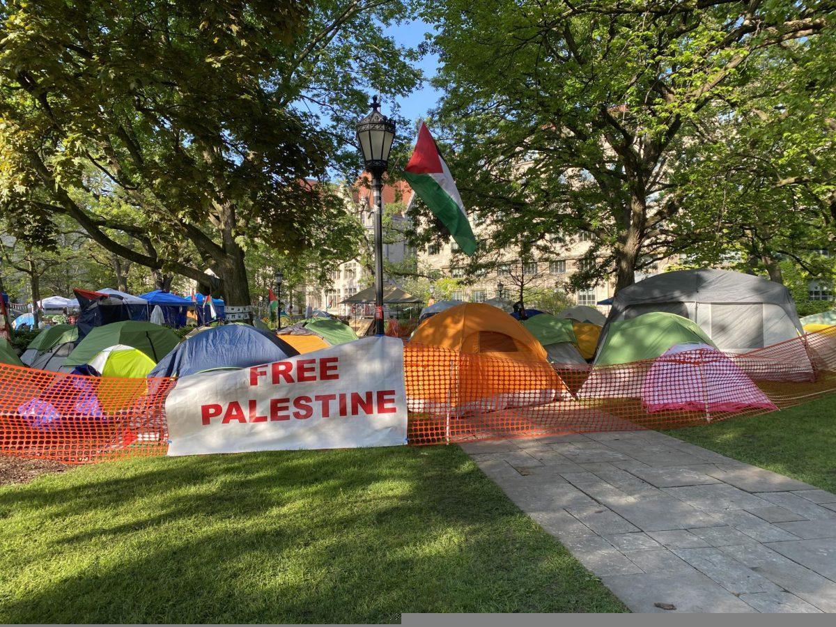 Various tents are pitched on the campus of University of Chicago, behind plastic orange construction fencing and a sign reading "Free Palestine"