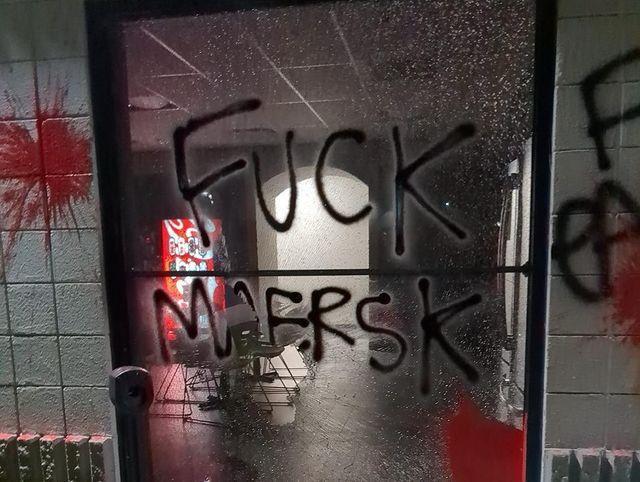 A shattered window with a dimly-lit break room behind it is covered in paint and graffiti. "FUCK MAERSK" is written on the window, with various red splatters of paint around it.