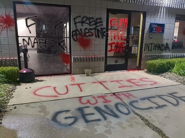 An office building entrance is vandalized. A broken window has "FUCK MAERSK" spray-painted on it. Other messages are spray-painted on the wall, door and sidewalk: "FREE PALESTINE", "FUCK THE IOF", "INTIFADA" and "CUT TIES WITH GENOCIDE". Splatters of red paint are all around.