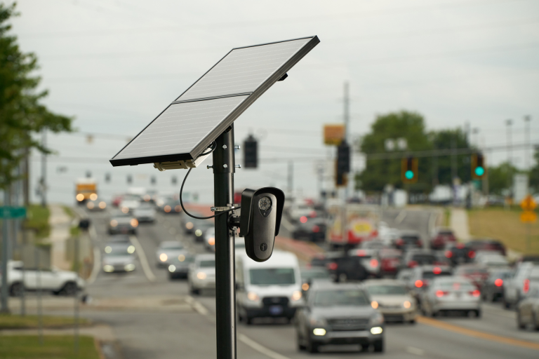 File photo of a Flock Camera installation. A solar panel sits atop a pole, with a cable connecting it to an oblong, black camera mounted lower on the pole.