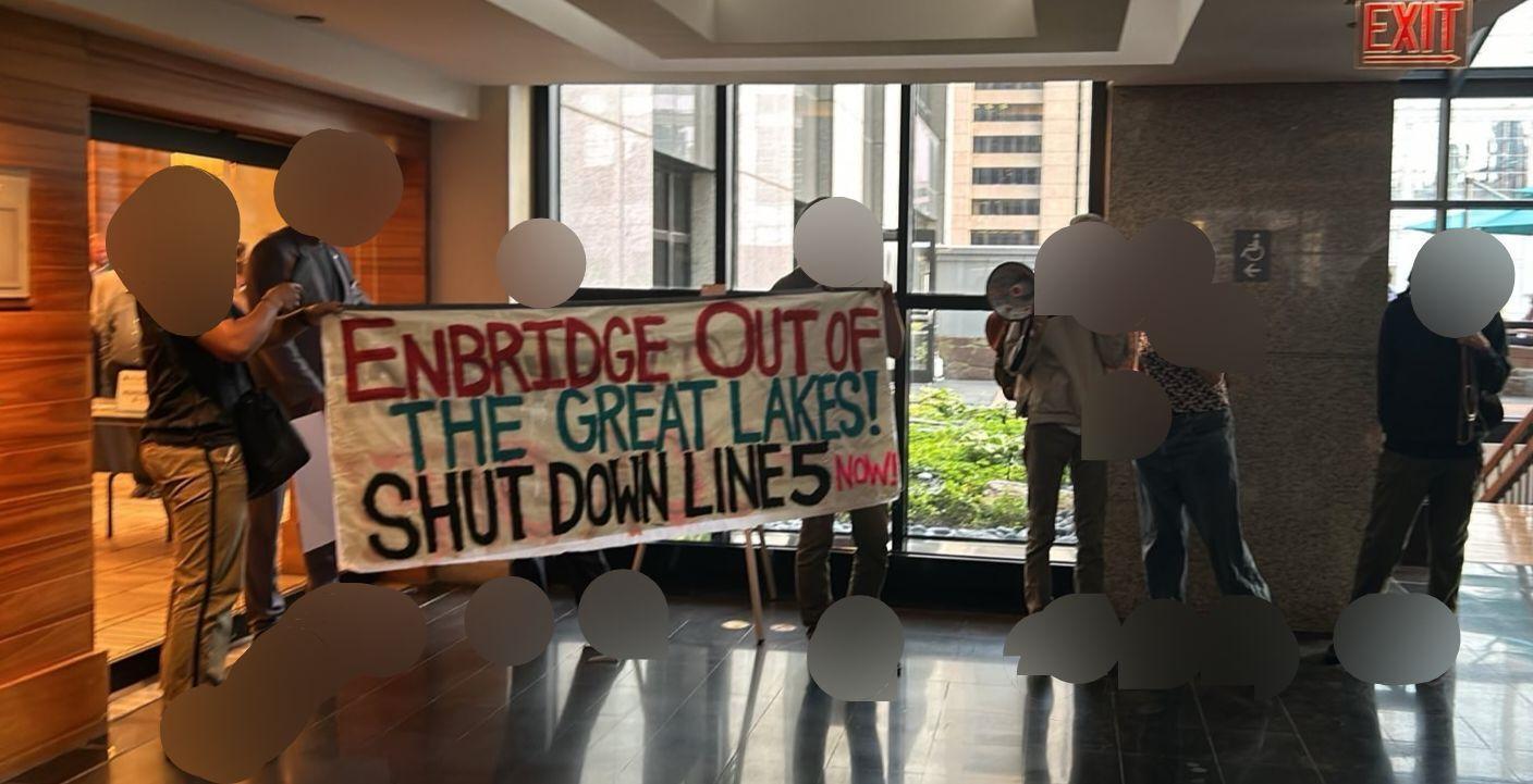 A group of people hold a banner somewhere inside a hotel conference area. The banner reads "Enbridge out of the great lakes! Shut down line 5 now!"