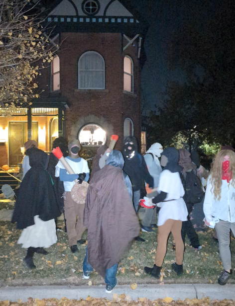 A crowd in peasant attire stand in front of a large house at night.