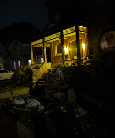 The front porch of a large house at night with the porch lights on. Prop gavestones are visible in the yard with caution tape, toilet paper, and various other devil's night materials and trash.