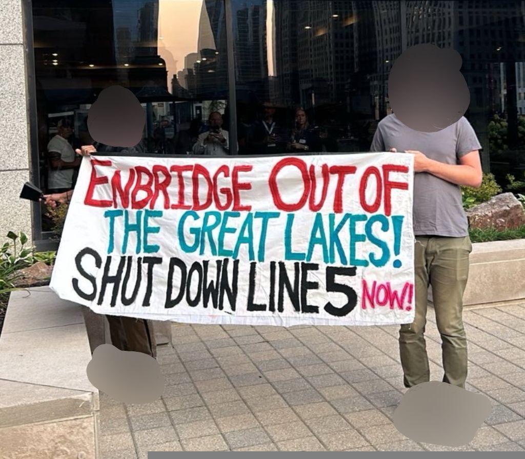 Two people hold a banner reading "Enbridge out of the great lakes! Shut down line 5 now!" in front of a hotel