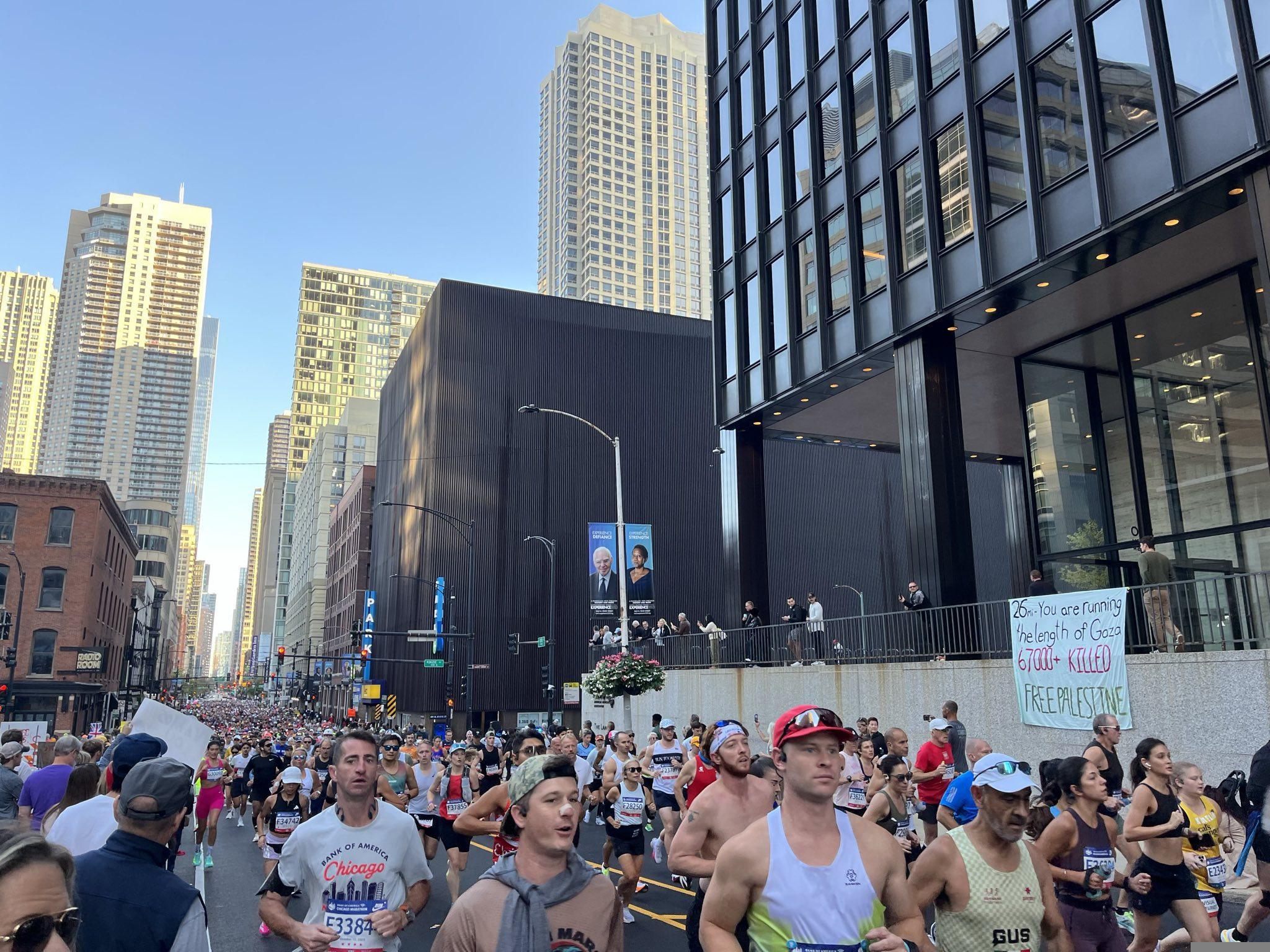 Marathon participants run down a street in downtown Chicago, past a hand-painted banner which reads "26mi - you are running the length of Gaza. 67000+ KILLED. FREE PALESTINE."