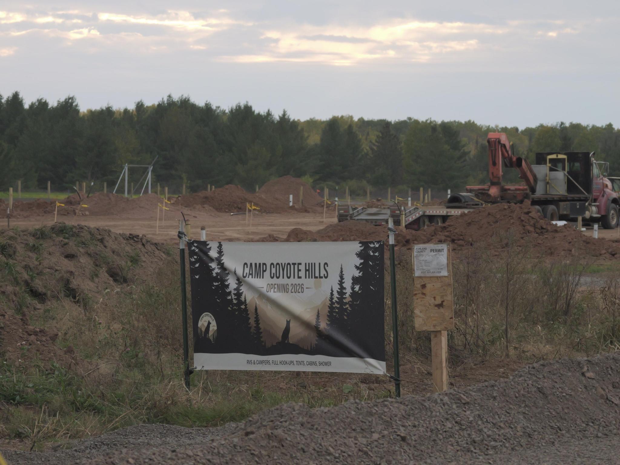 A large open field with survey stakes, exposed earth and construction equipment. A banner alongside the driveway into the worksite reads "Camp Coyote Hillls" and "Opening 2026"