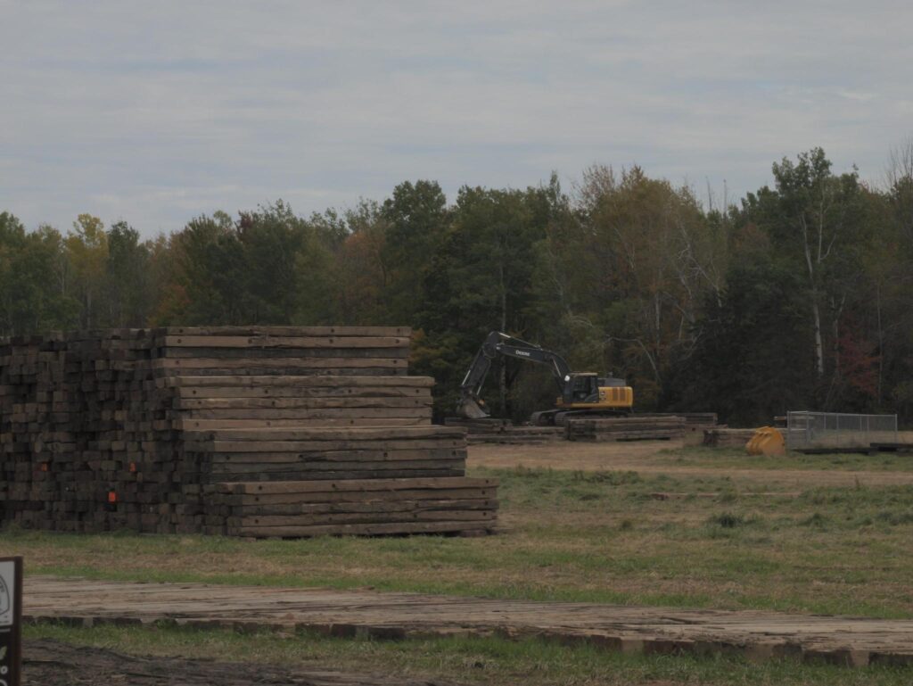 An open field with trees in the background. There is construction equipment in the field, and tall piles of square timber in the foreground, like railroad ties.