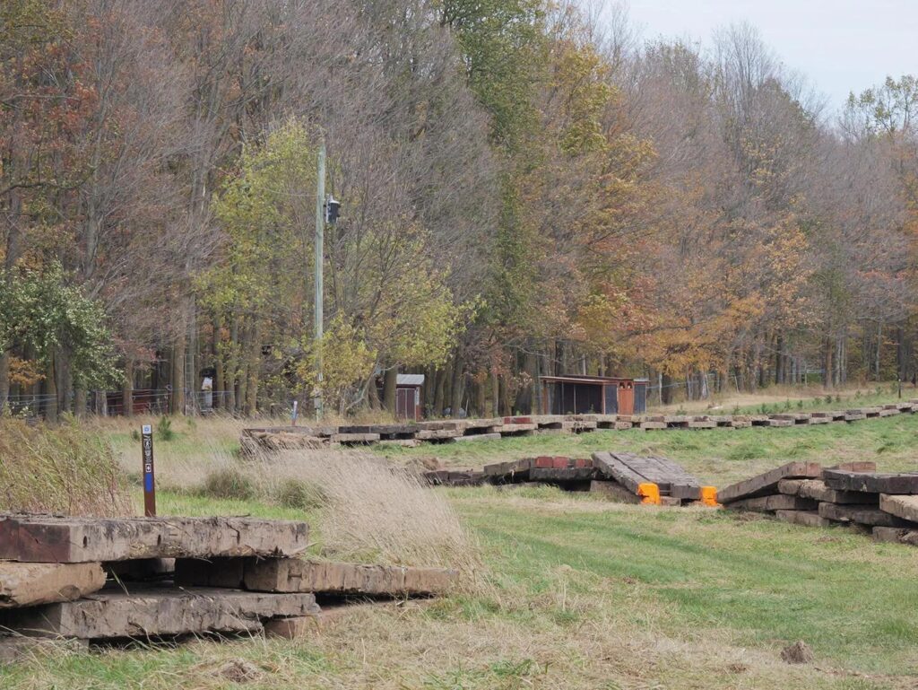 A trail along the edge of an open field, with trees on the left side. The trail is divided from the field by short stacks of square timber, like a wide fence.